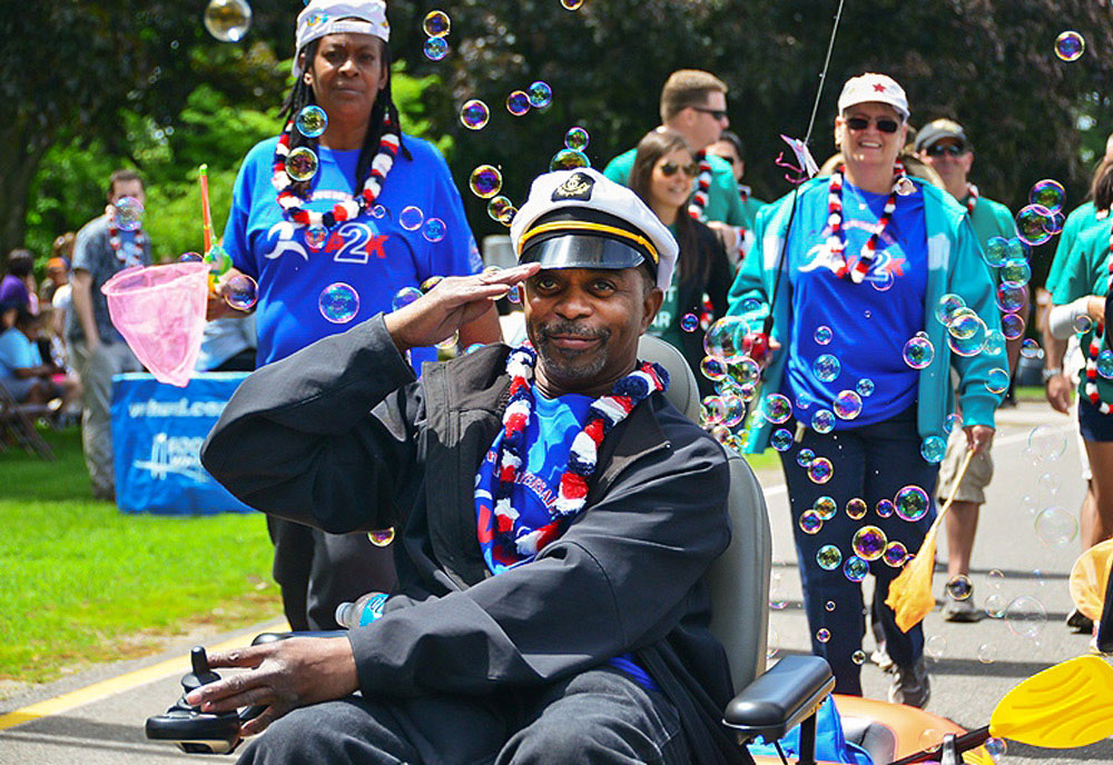 A Veteran wearing a sea captain's hat salutes while rolling his motorized wheelchair among others walking around him