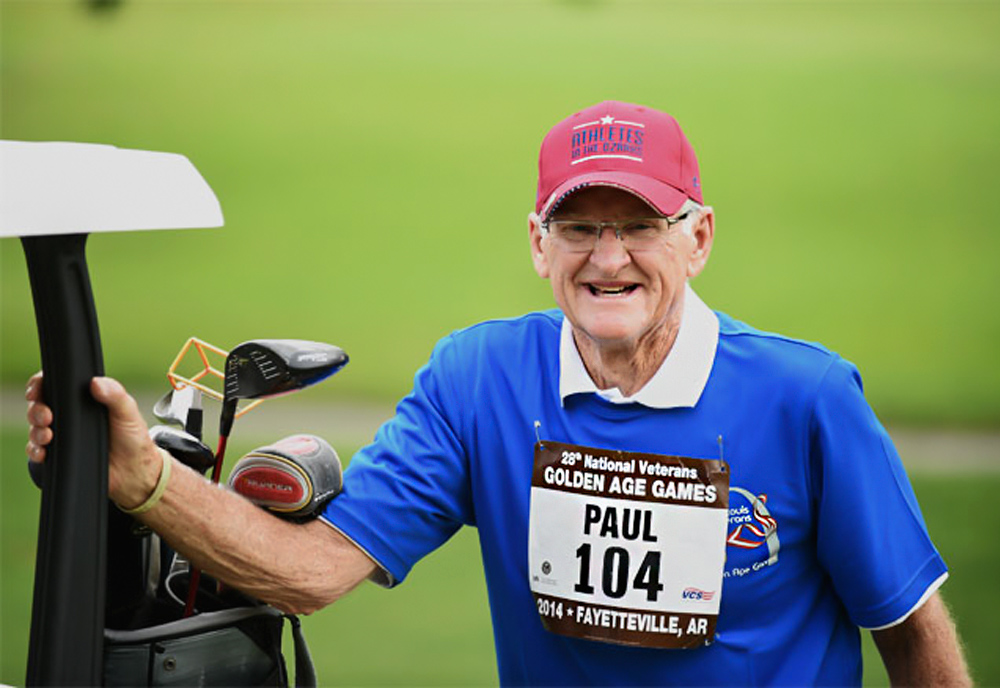 An older man with a sporting competitor's numbered bib on his shirt leans on a golf cart carrying his clubs on a golf course