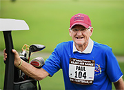An older man with a sporting competitor's numbered bib on his shirt leans on a golf cart carrying his clubs on a golf course