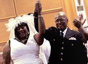A man and woman dressed in wedding attire smile and hold hands above their heads