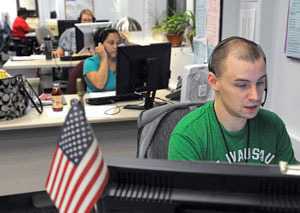 Several people sit at computers at their desks, speaking on headsets with a small American flag in the foreground