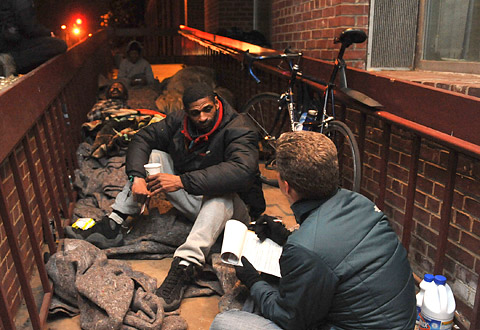 a man with a notepad talks with a homeless man on the street at night