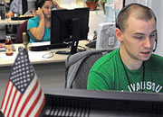 A man and woman sit at computers at their desks, speaking on headsets with a small American flag in the foreground