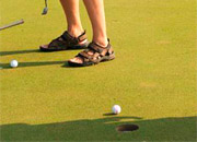 Feet in sandals stand on a golf green as a golf ball nears the hole