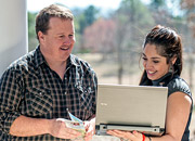 A man and woman stand outside, smiling and looking at a laptop