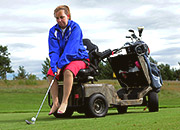 A disabled woman sits on the back of a golf cart, preparing to swing a club