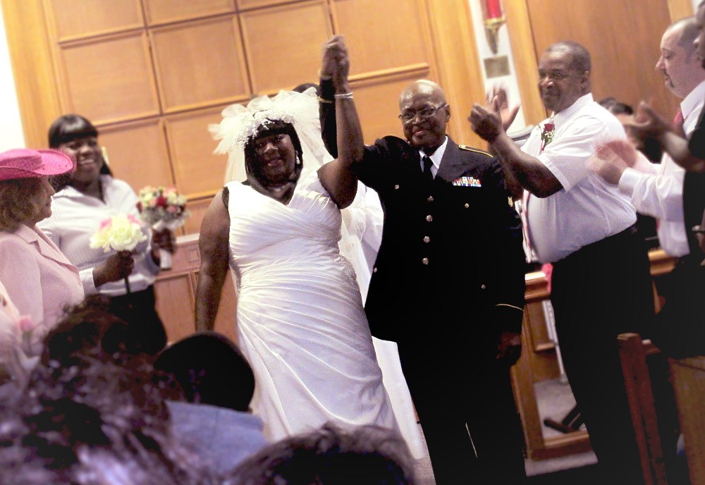 A man and woman dressed in wedding attire smile and hold hands above their heads, surrounded by people clapping