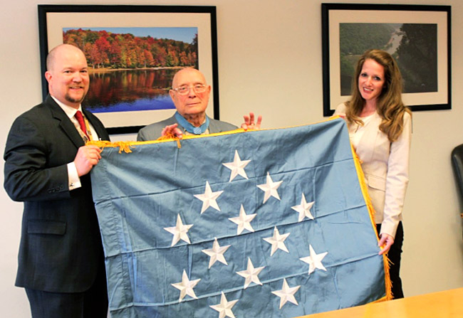 Veteran stands with a man and woman holding a Medal of Honor flag