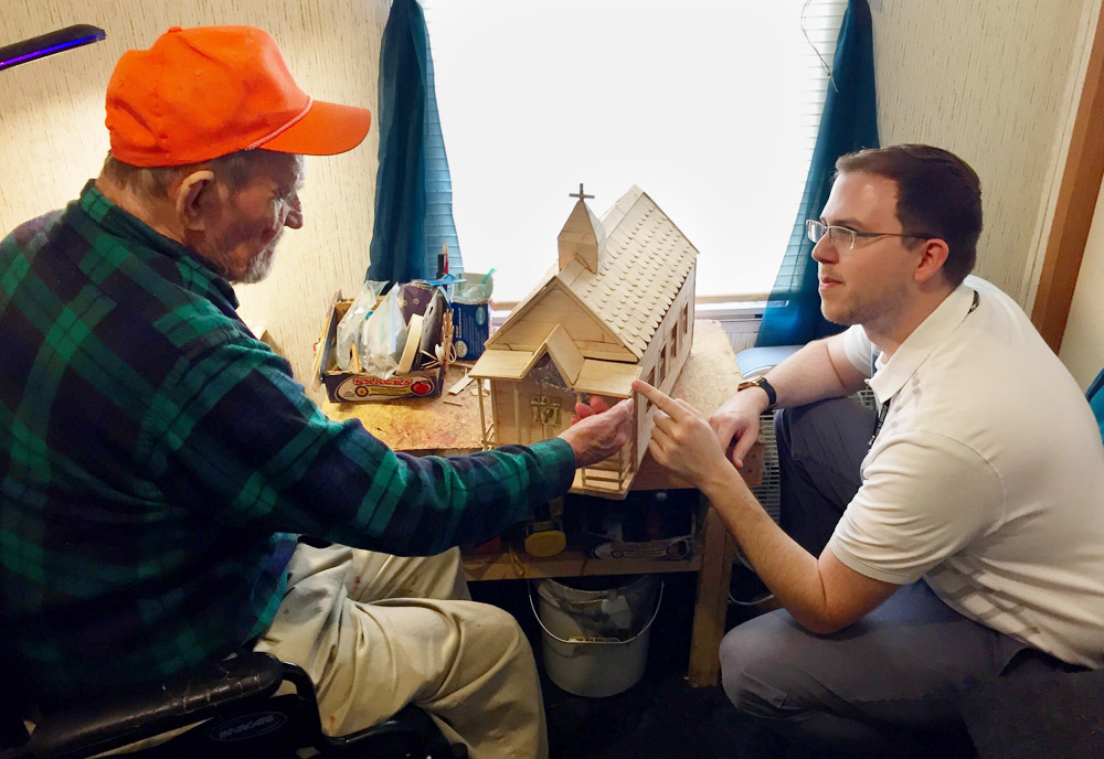 A man in a wheelchair shows a wooden dollhouse to another man. 