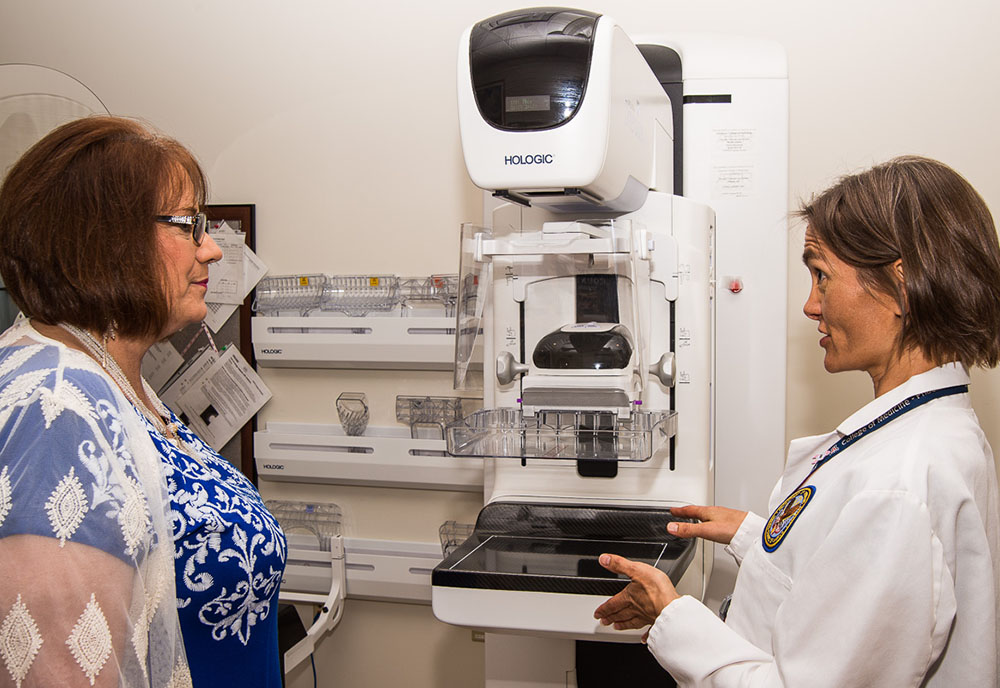 A woman in a lab coat talks to a female patient next to a breast imaging machine
