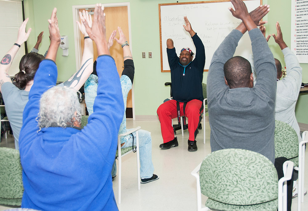 A man leads a seated classroom of participants facing him, practicing Tai Chi arm movements