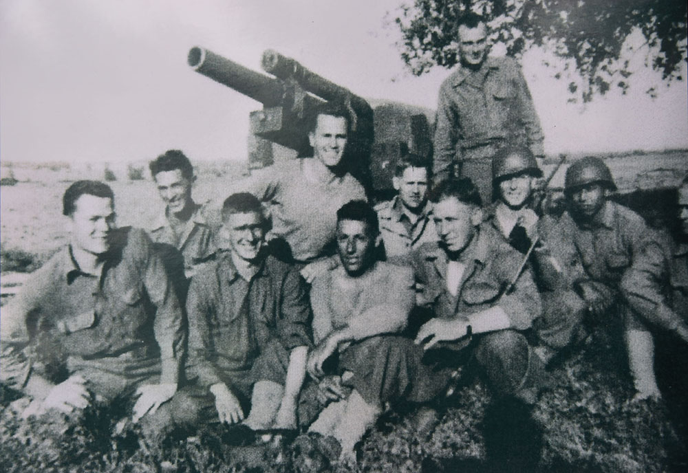 Ten men in Army field uniforms posing in front of a cannon