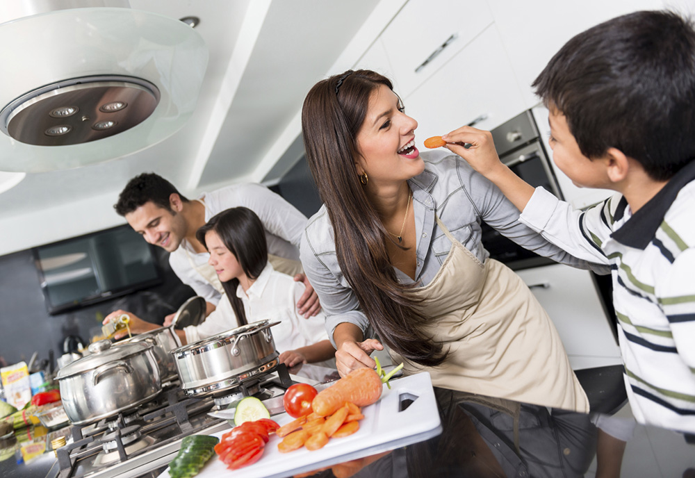 A mom and dad with a son and daughter cooking and sampling food in a kitchen