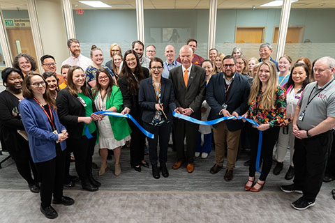 Pictured is a large group of health professionals standing in front of a blue ribbon. Dr. Ezgi Tiryaki is centered, holding scissors and just made the cut
