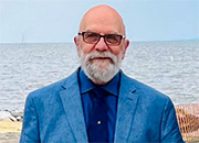 Man with white beard in blue suit standing on beach with ocean in background