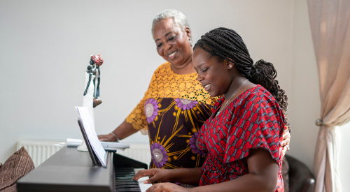 Older woman standing watching young woman seated playing piano 