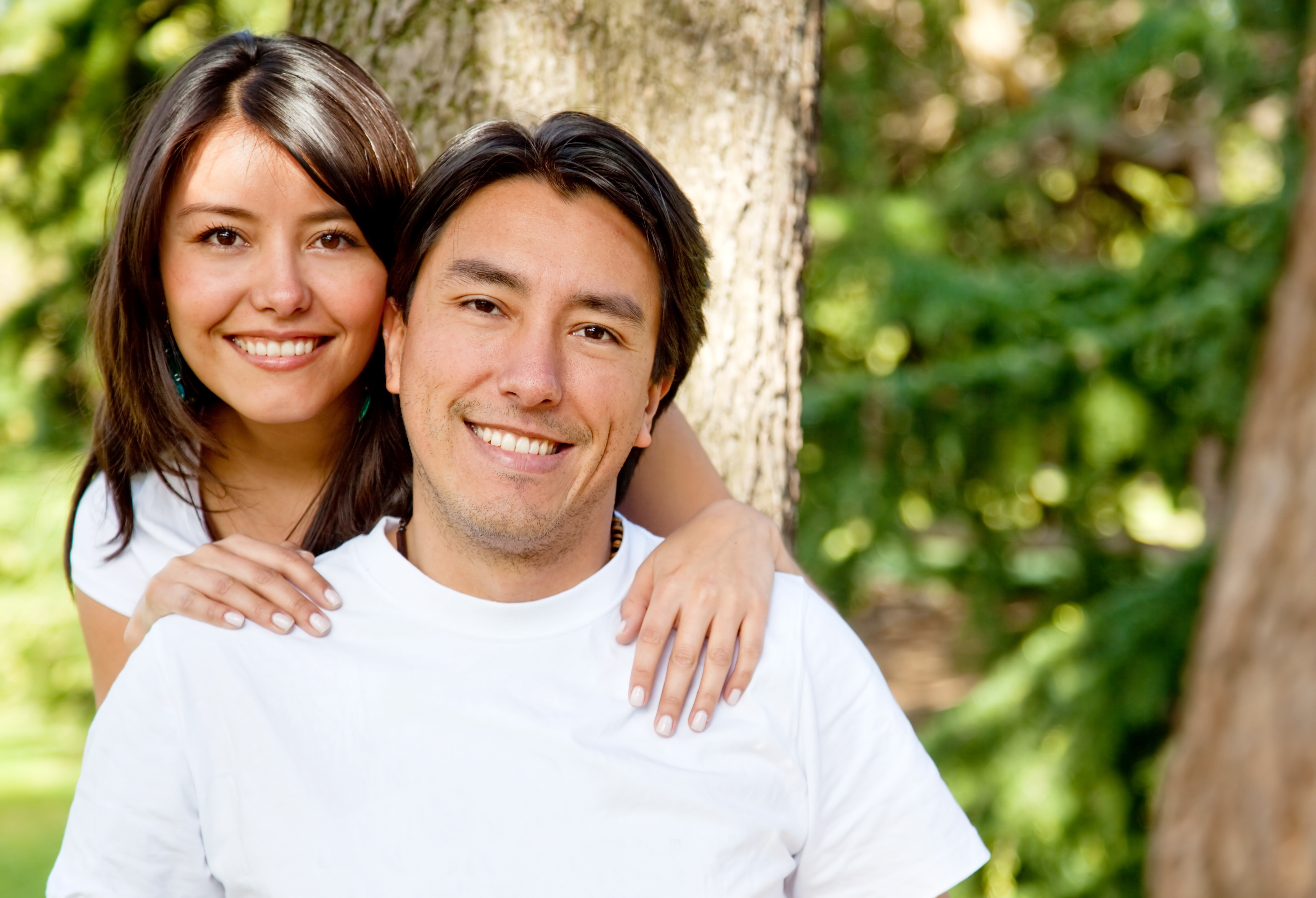 Close-up of a young man and woman both in white t-shirts, smiling, outside in a park.