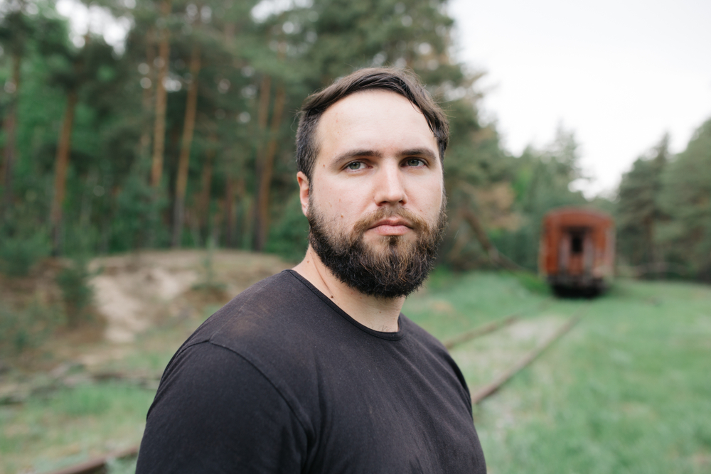 Young man with a thick beard and serious expression wearing a black t-shirt. The man is standing in a wooded area next to train tracks with a cabose on the tracks in the background.