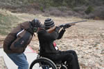 A man in a wheelchair fires a shotgun at an outdoor firing range.