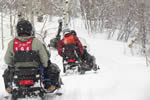 People on snowmobiles travel on a snow covered trail.