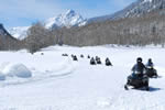 People on snowmobiles traveling on a snowcovered trail with mountains visible in the distance..