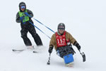 A man in a monoski is assisted from behind by an instructor as they ski down the mountain.