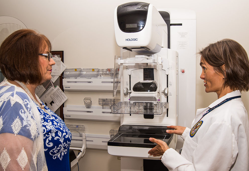 A woman in a lab coat talks to a female patient next to a breast imaging machine A woman in a lab coat talks to a female patient next to a breast imaging machine